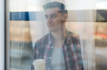 Handsome smiling man guy in plaid shirt looking through the window with a cup of morning coffee...