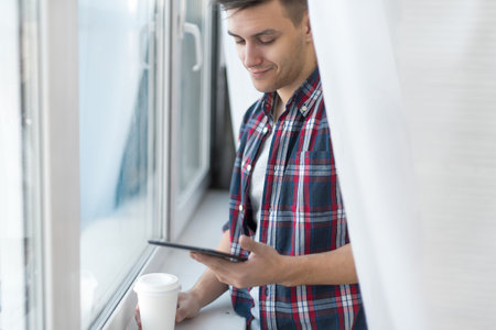 Handsome smiling man guy in plaid shirt near window with a cup of morning coffee and tablet.
