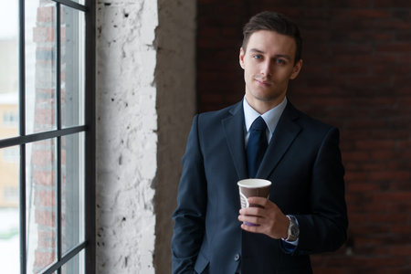 Businessman drinking coffee in office standing near window looking at camera