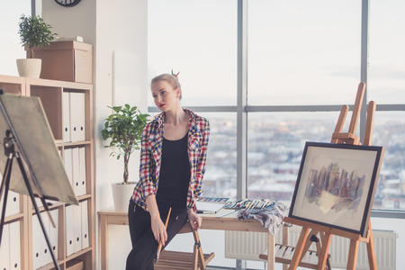Female painter standing in workshop, watching at drawing attentively, holding paintbrush. Artist looking on picture concentrated, front view portraitの写真素材