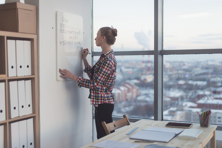 Businesswoman writing day plan on white magnet board, modern office. Side view of caucasian female employee planning schedule in morning at workplaceの写真素材