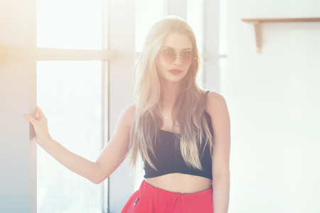 Close-up portrait of young beautiful woman wearing glamorous romantic summer look, with sunglasses in bright morning light, against large windowの写真素材