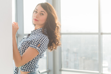 Beautiful woman with long loose hair standing next to wall in spacious light room, posing, looking at cameraの写真素材