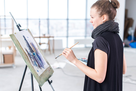 Female painter drawing in art studio using easel. Portrait of a young woman painting with aquarelle paints on white canvas, side view portrait.の写真素材