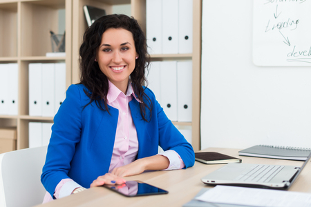 Portrait of a woman sitting in office, smiling, looking at camera. Young confident female business worker ready for the work dayの写真素材