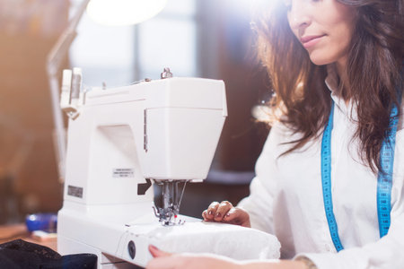 Close-up view of sewing machine and young female tailor working on it while sitting in a workshop