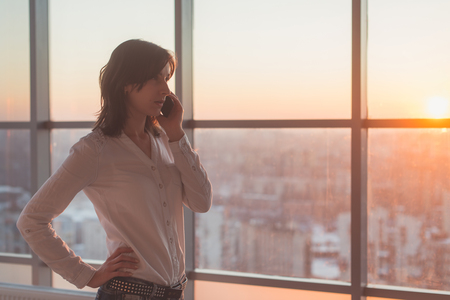 Young woman talking using cell phone at office in the evening. Female businesswoman concentrated, looking forward, calling by mobile device on her workplace.の写真素材