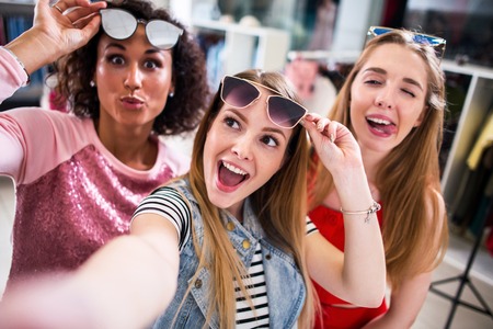 Three female friends taking selfie making faces raising sunglasses in clothing and accessories outletの写真素材
