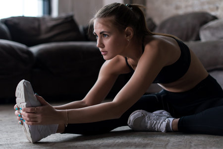 Slim sporty Caucasian girl sitting on floor doing stretching exercise indoors