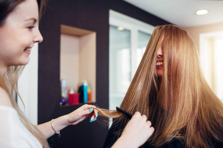 Female customer with long straight fair hair laughing while a hairdresser combing out strands in front of her face working in beauty salon.の写真素材