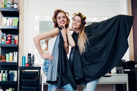 Portrait of two pretty blondes wearing hair rollers and cape standing in funny silly pose looking in camera at hairdressing and beauty studioの写真素材