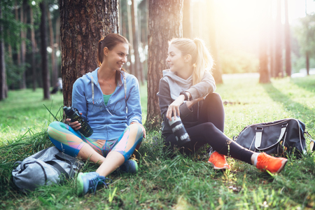 Two young women in sportswear sitting under trees in the forest drinking water, talking and resting after training outdoorsの写真素材
