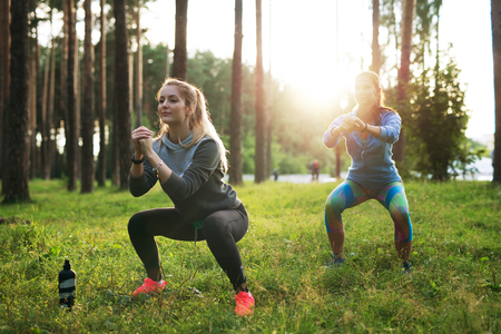 Fit young Caucasian women wearing jumpsuits working out in the forest doing squats in the early morningの写真素材