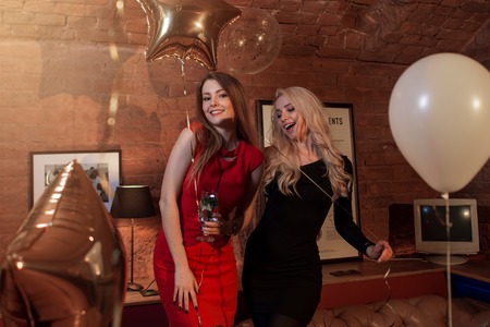 Two pretty women in cocktail dresses posing with balloons at birthday party in stylish cafeの写真素材