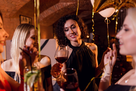 Close-up shot of positive beautiful female friends raising glasses of wine to happy event sitting in fashionable restaurantの写真素材