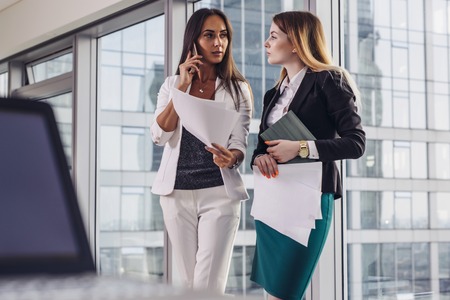 Two businesswomen holding documents talking to their customer by mobile phone offering different solutions standing in the fashionable officeの写真素材