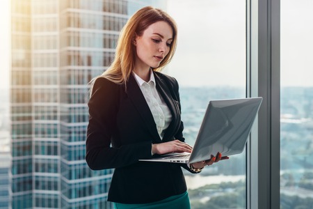 Young female lawyer working in her luxurious office holding a laptop standing against panoramic window with a view on business districtの写真素材