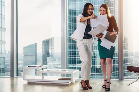 Female businesswomen wearing formal outfit discussing documents standing in office hallwayの写真素材