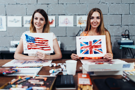 Two smiling female designers of prints showing their works, American and British flags drawn with watercolor technique, sitting at their work desk in creative officeの写真素材