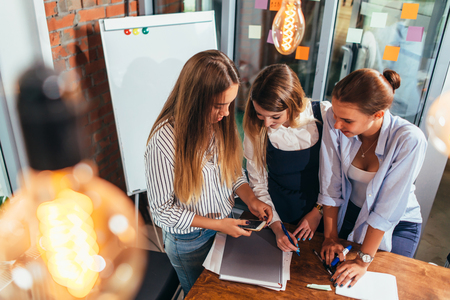 Top view of three cheerful female students looking at mobile phone screen standing in classroomの写真素材
