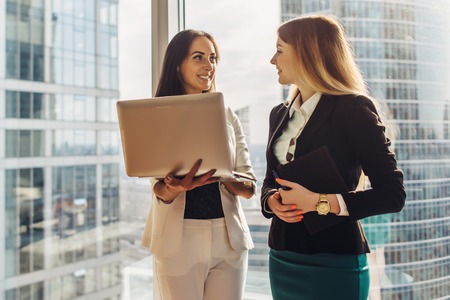 Smiling young women with laptop standing and talking in officeの写真素材