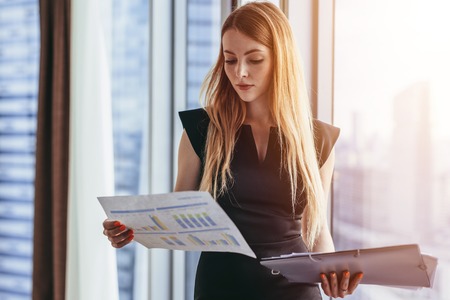 Female financial analyst holding papers studying documents standing against window with city viewの写真素材