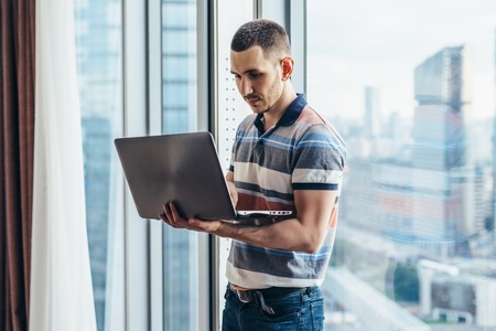 Businessman holding a laptop working standing in office near the window with city viewの写真素材