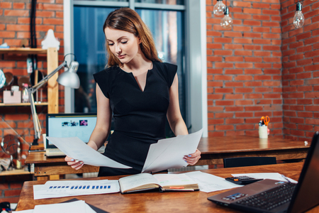 Serious woman reading papers studying resumes standing at work desk in stylish officeの写真素材