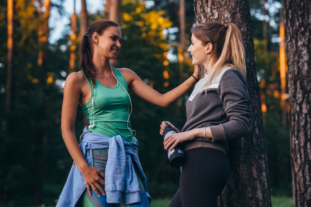 Two female runners relaxing after workout standing near the tree talking in parkの写真素材