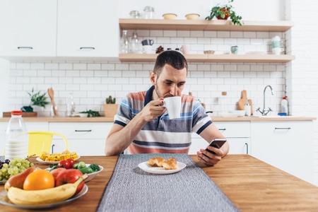Young man sitting at table in the kitchen with a cup of coffee using mobile phone.の写真素材