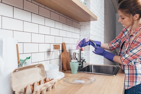 Young woman wearing glove cleaning sink, wiping faucet in kitchenの写真素材