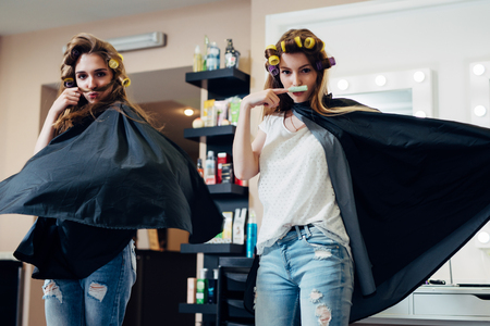Two female friends goofing around in front of the camera making moustache from hair and curler standing in flying cape like heroes at beauty shopの写真素材
