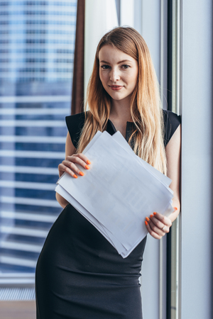 Portrait of pretty smiling young woman holding papers standing at window with cityscape viewの写真素材