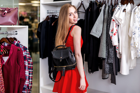 Side view of pretty girl wearing red set doing shopping alone choosing, holding, examining new bag in fashion storeの写真素材