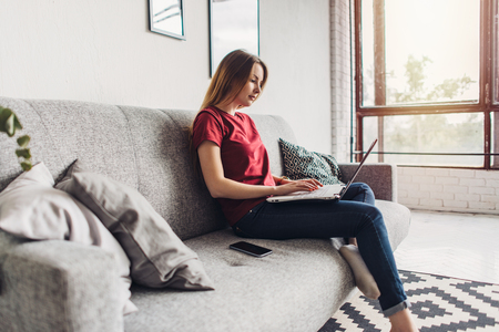 Beautiful young woman sitting on sofa and using laptop at homeの写真素材