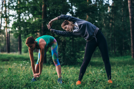 Two female runners doing warming-up bending exercises stretching back and leg muscles before running in parkの写真素材