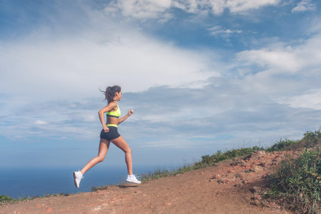 Athletic woman running up the mountain with sky and sea in background. Professional runner doing...