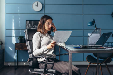 Woman in office talking on mobile phone holding document working at her workplace.の写真素材