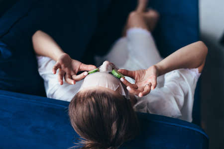Woman lies on a couch with a sheet mask over her face and cucumber slicesの写真素材