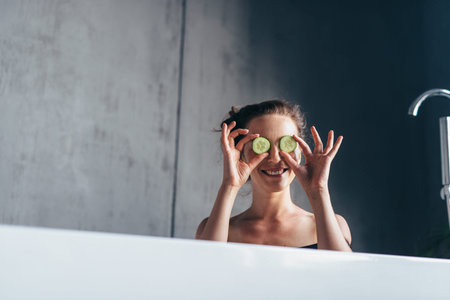 Cheerful woman sits in the bathtub and holds slices of cucumber in front of her face