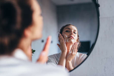 Woman washes her face in front of the mirror, applying foam to her cheeksの写真素材
