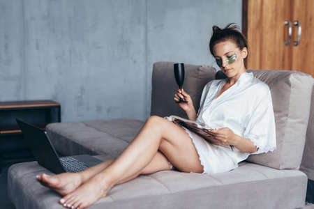 Young woman sitting on sofa with patches under eyes and reading magazineの写真素材