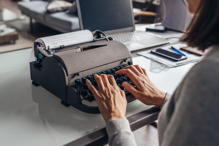 Woman author composes text on a typewriter sitting in her working room.
