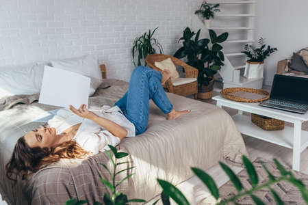 Young woman is lying on her bed at home reading documentsの写真素材