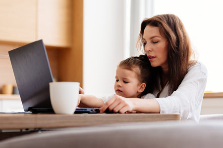 A woman works at a kitchen table with a laptop and a childの写真素材