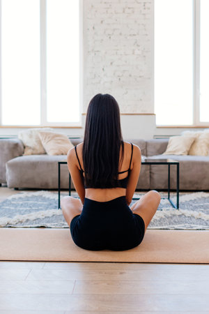 Woman sitting on floor in meditative pose her legs crossed. She is facing away from camera, wearing black workout clothing sports bra and shortsの写真素材