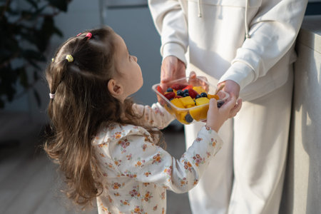 Mother and child in home kitchen, girl takes fruit from container