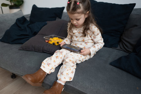 Girl sits alone on sofa at home, eating fruit and watching smartphoneの写真素材
