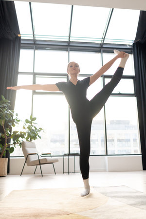 A young woman in sportswear practices a standing yoga pose indoors, raising one leg high and holding...