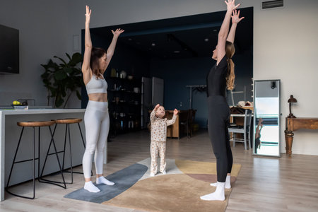 Two women in sportswear and a child practicing yoga together at home, standing with arms raised...